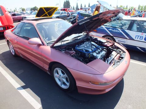 2003 Ford Crown Victoria - salmon-colored sports coupe with raised hood exposing a blue-finished engine, parked at an outdoor car show.