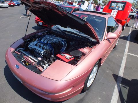 2003 Ford Crown Victoria - mauve-pink Ford sports coupe sits at an outdoor car show with its hood up, exposing a transverse V6 engine bay.