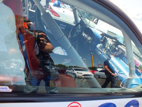 2003 Ford Crown Victoria - side window of a race car reflects the photographer and the car-show lot, with a Momo steering wheel visible inside.