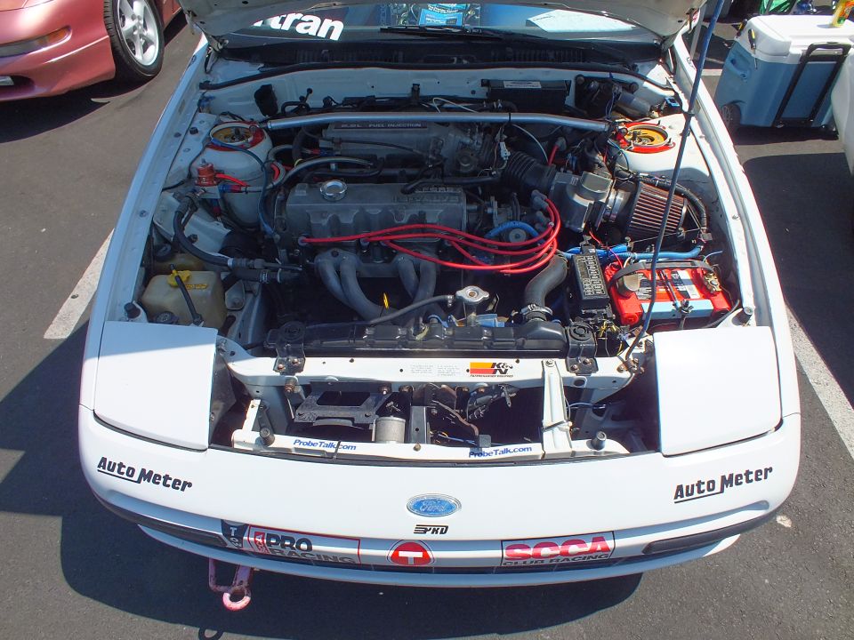 Open engine bay of a white car showing a four-cylinder engine with red plug wires and decals