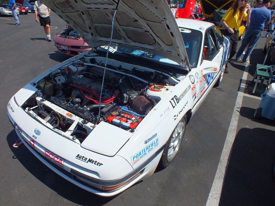 White race-liveried sports coupe with open hood and sponsor decals at an outdoor car show