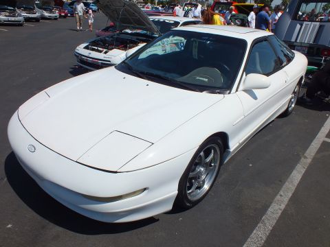 2003 Ford Crown Victoria - clean white sports coupe with a rounded nose and five-spoke wheels sits parked among show cars on asphalt.