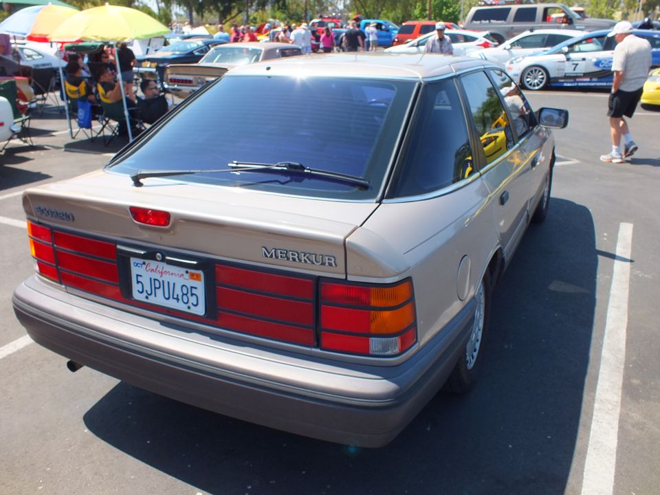 Rear of a tan Merkur hatchback with California plate at a sunny car show
