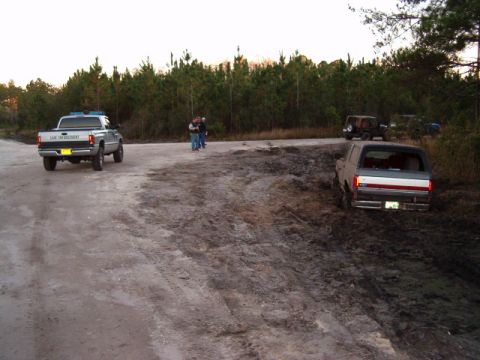 1991 Ford Bronco - A gray Ford Bronco is stuck in the mud off a dirt road, with other vehicles and people in the background.
