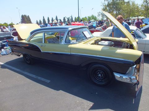 2003 Ford Crown Victoria - two-tone yellow-over-black 1950s American sedan sits with its hood up in a sunny car show parking lot full of spectators.