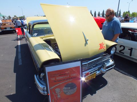 2003 Ford Crown Victoria - pale yellow 1950s car displays its raised hood beside a framed poster titled The Bonneville Story at an outdoor show.