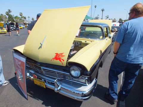 2003 Ford Crown Victoria - pale yellow 1950s sedan with chrome grille shows a red flying-horse Pegasus emblem on its raised hood as a spectator looks on.