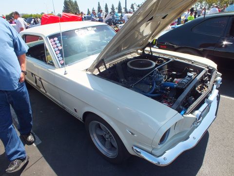2003 Ford Crown Victoria - white 1960s fastback muscle car sits with hood raised, revealing a chrome air cleaner, at a crowded outdoor car show.