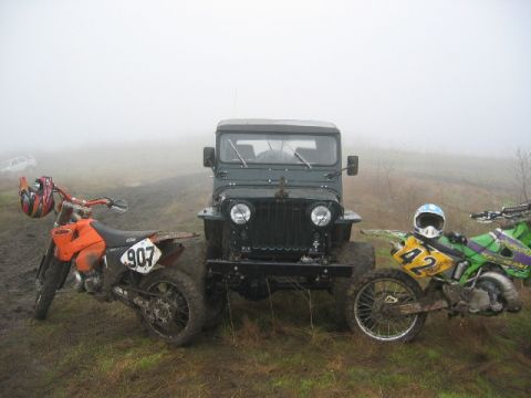 1989 Ford Bronco - A green jeep is flanked by two dirt bikes, one orange and one green, in a misty outdoor setting.