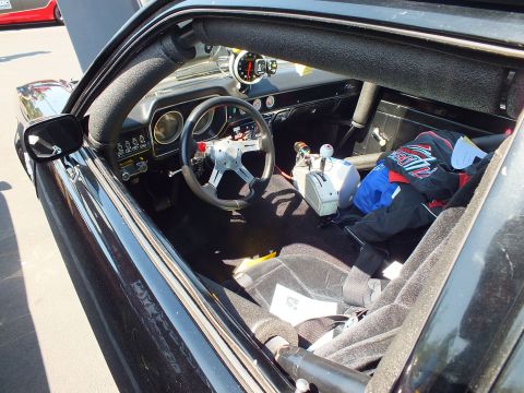 2003 Ford Crown Victoria - stripped interior of a black race car shows a roll cage, dash gauges, racing steering wheel and gear piled on the floor.