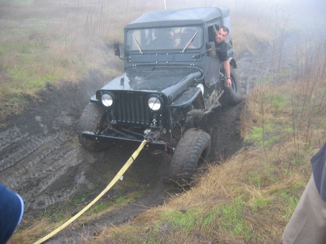 Dark vintage flat-fender 4x4 in deep mud with yellow tow strap, man leaning out