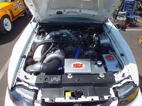 Engine bay of white Mustang showing engine with RSCA placard and intake piping