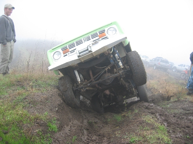 Green and white SUV stuck nose-down in a muddy off-road rut, front axle exposed