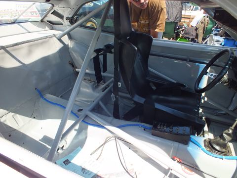 Stripped race car interior with roll cage, single black bucket seat, harness and gear shifter