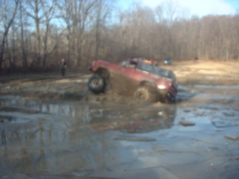 1993 Ford Bronco - A red SUV is stuck in a muddy area with trees in the background and another vehicle visible.