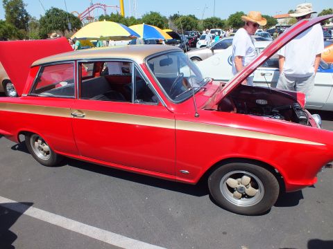 Red two-door sedan with gold side stripe and open hood at an outdoor car show