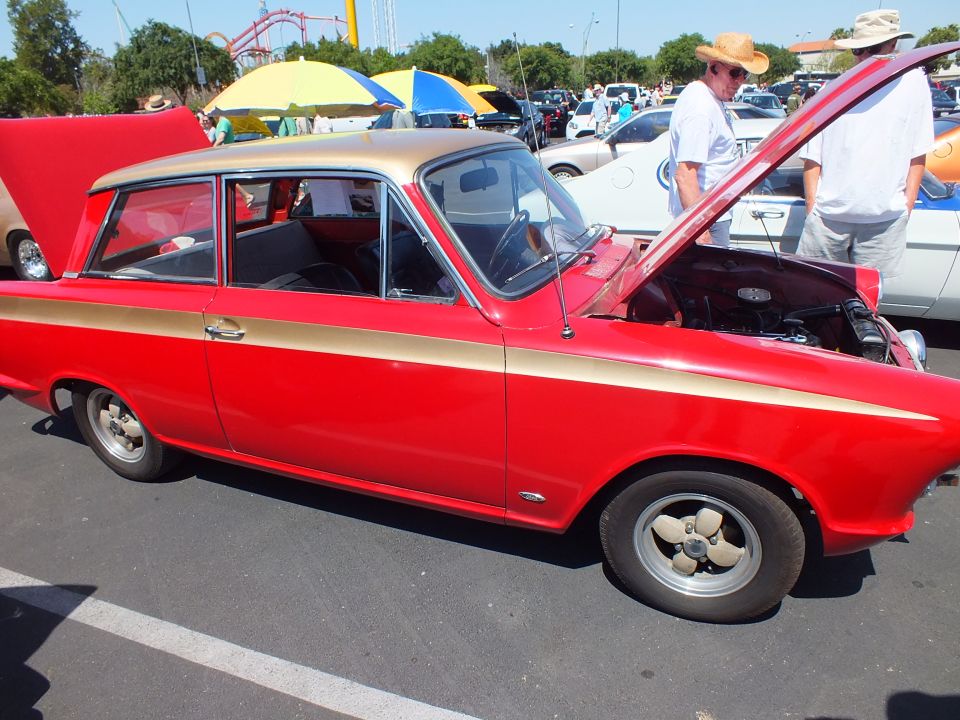 Red two-door sedan with gold side stripe and open hood at an outdoor car show