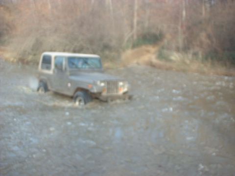 1993 Ford Bronco - A gray Jeep Wrangler navigates through a shallow body of water, kicking up spray.