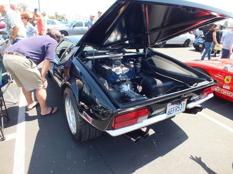 Black mid-engine coupe at car show with rear hatch up, spectators nearby, plate 6EFR537