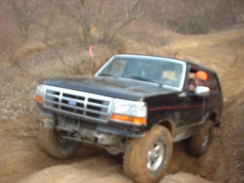 1993 Ford Bronco - Black full-size Ford Bronco with a chrome grille climbs a muddy off-road berm on tall mud tires caked with mud, a person in an orange cap l…