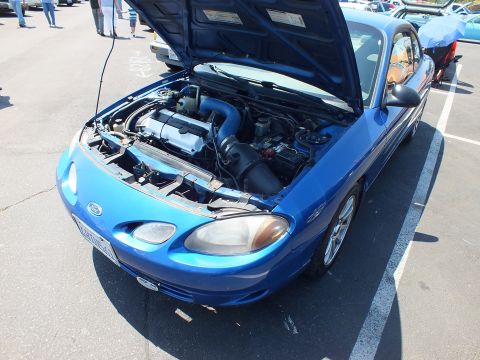 Blue Ford coupe with hood up showing turbocharged engine at car show