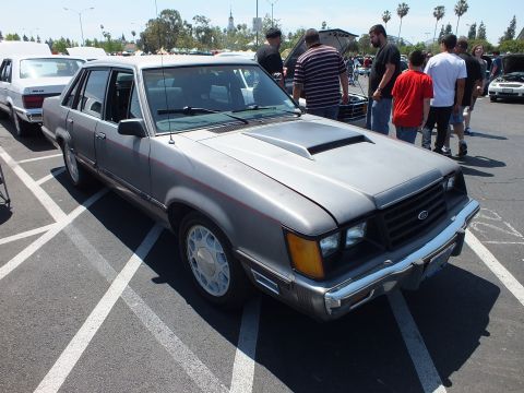 Silver Ford four-door sedan with hood scoop at sunny car show