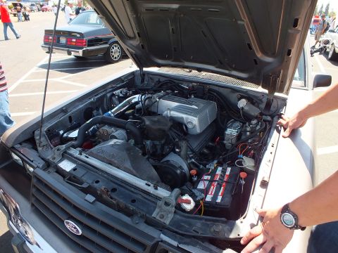Open engine bay of a car at a show with a person's hand resting on the fender