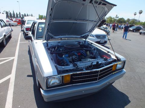Front of a silver 1980s coupe with hood up showing engine at a car show