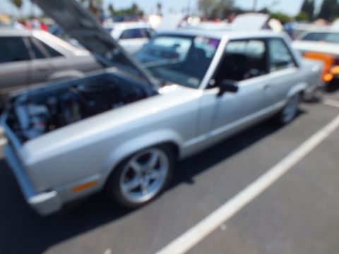 Blurry side view of a silver two-door car with hood up at a car show