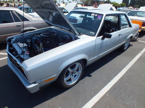 Silver two-door coupe with hood up and aftermarket wheels at a car show