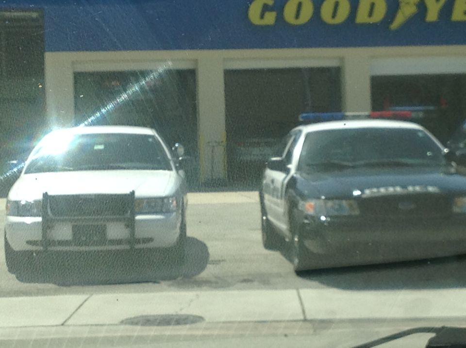 White Crown Victoria with push bar parked next to a black-and-white police Crown Victoria at a Goodyear shop