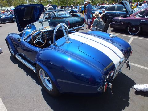 Rear three-quarter view of a blue Cobra-style roadster with white stripes, hood open