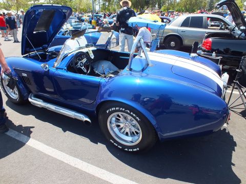 Rear three-quarter view of a blue Cobra-style roadster with white stripes on Firehawk tires