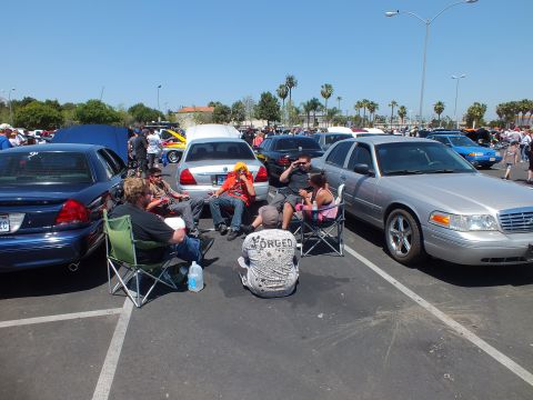 People in folding chairs sit between parked cars at an outdoor car show