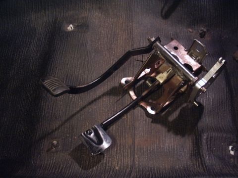 1991 Ford LTD Crown Victoria - A removed brake pedal and parking-brake pedal assembly laid on a bare, stripped floor pan, showing the black pedal arms, ribbed rubber brak…