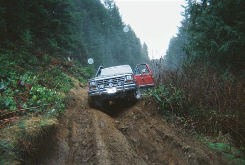 1983 Ford Bronco - A red and black Ford truck navigates a challenging muddy incline on a forest trail.