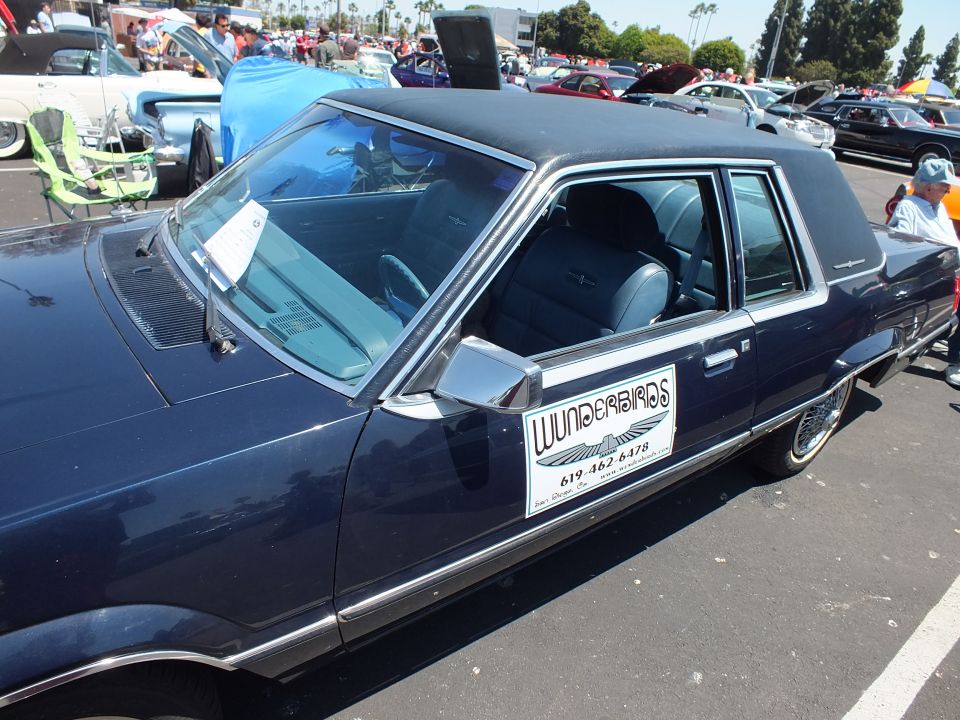 Dark blue two-door Ford at an outdoor car show with vendor sign in window
