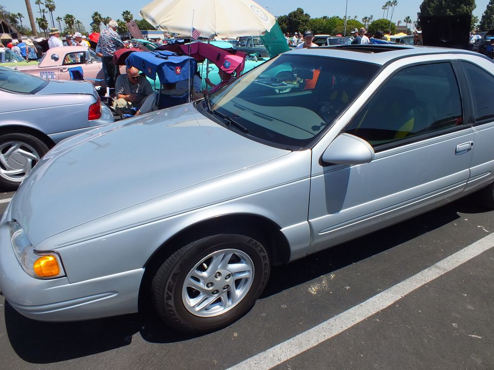 Silver two-door coupe parked at an outdoor car show with vendor tents