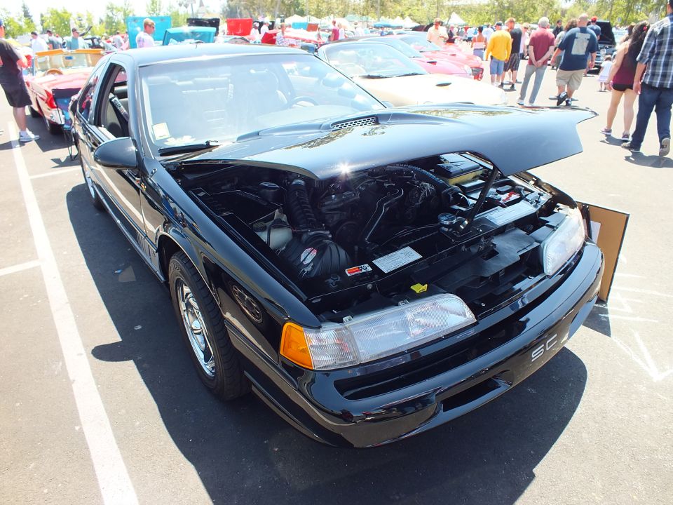 Black coupe with 'SC' badge and open hood at a sunny car show