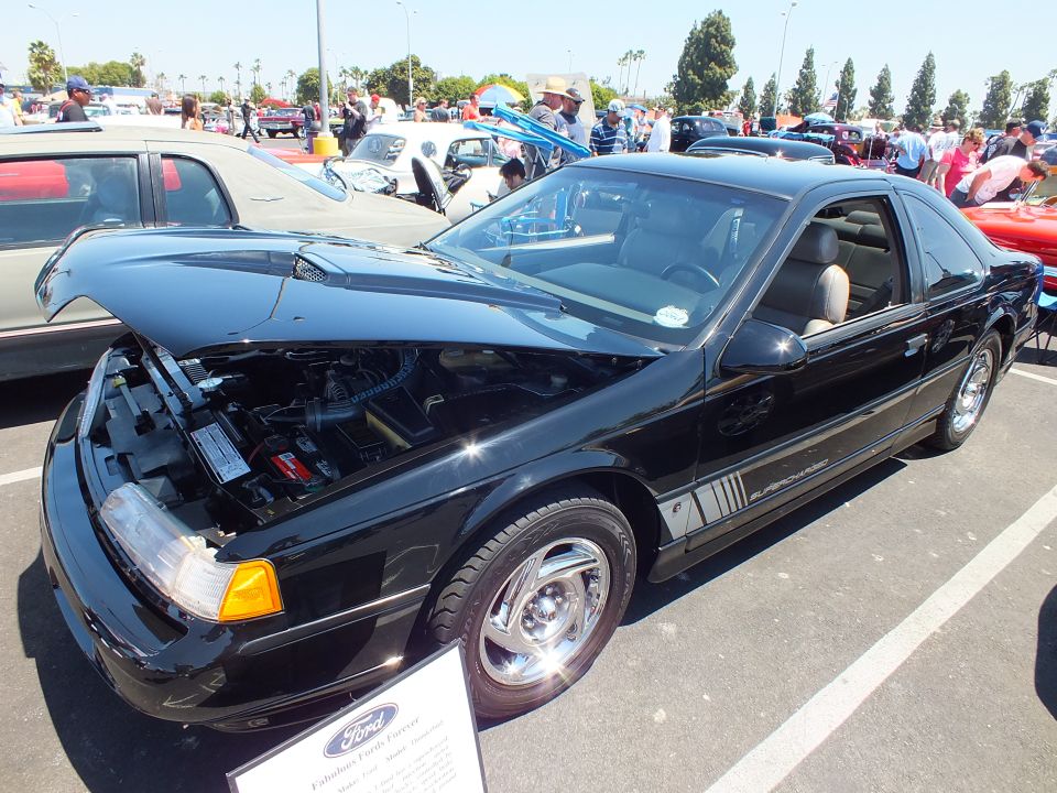 Black coupe with open hood and Ford placard at an outdoor car show
