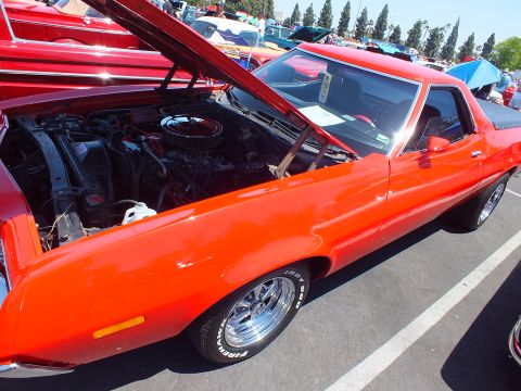 Orange-red car-based pickup with hood up and chrome wheels at show