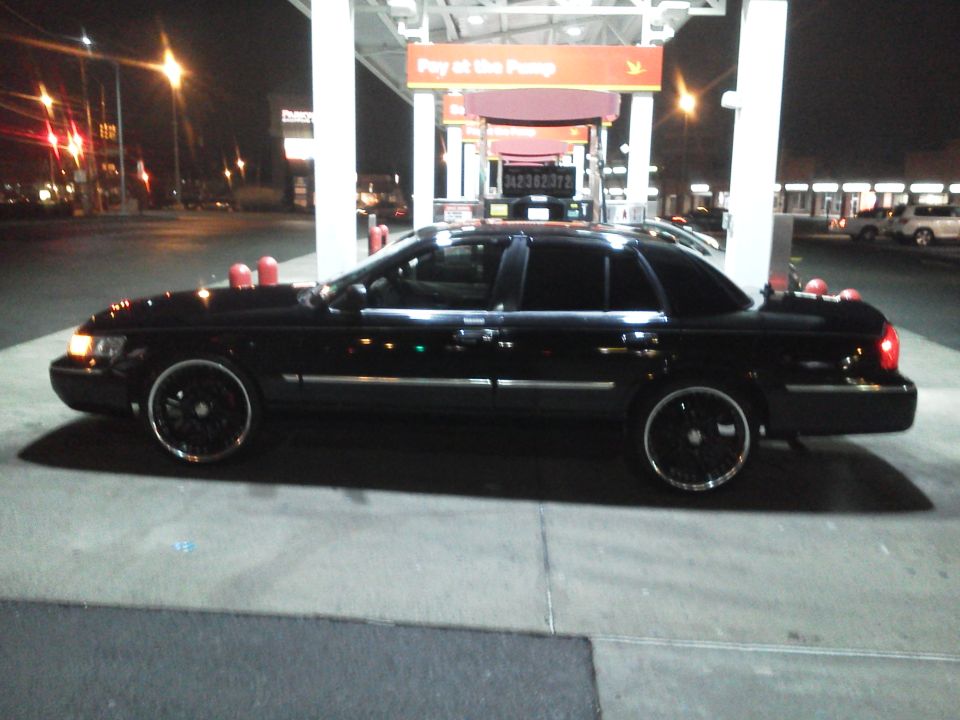 Black full-size sedan on large black rims at gas station pump at night