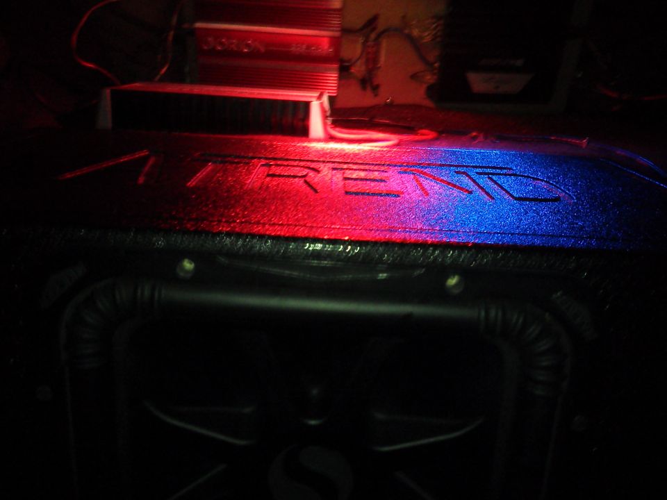 Car audio amplifier with red and blue lighting on a textured surface with 'TREND' embossed, above a subwoofer box