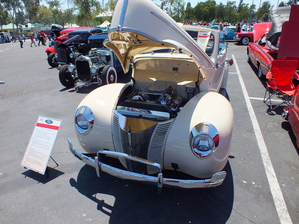 Cream 1940 Ford front view with open hood, chrome grille and Ford info placard at show