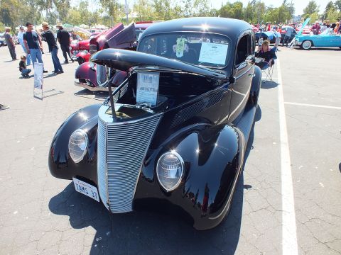 Black 1937 Ford coupe hot rod front view with open hood and chrome grille at car show