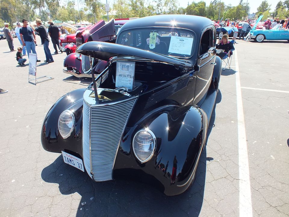 Black 1937 Ford coupe hot rod front view with open hood and chrome grille at car show