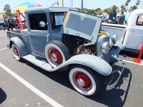 Gray 1930s Ford pickup hot rod with red-spoke whitewall wheels and open hood at car show