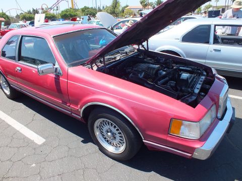 Red 1980s Lincoln Mark VII coupe with open hood and BBS-style wheels at car show