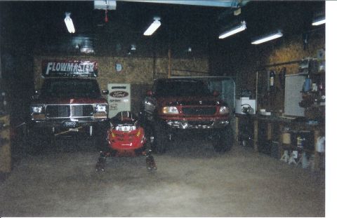 1978 Ford Bronco - Dim scanned photo of a home shop with two lifted red Ford trucks parked front-on, a square-body with a chrome grille and TORCO bumper plate…