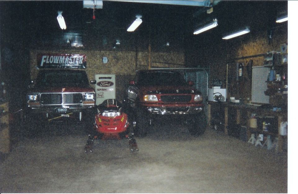 Two lifted red Ford trucks and a red snowmobile in a shop with a Flowmaster banner